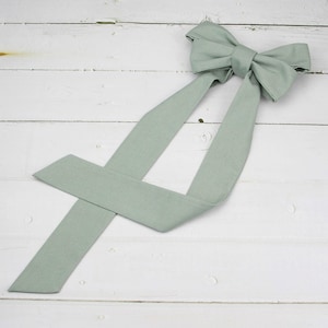 Può includere: Un fiocco verde salvia con lunghe code è appoggiato su una superficie di legno bianco. Il fiocco è legato in modo classico, con le code che si estendono diagonalmente. Il tessuto sembra morbido e strutturato, adatto per accessori per capelli o abbigliamento.