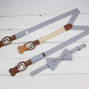 Può includere: Un set di bretelle grigio chiaro con dettagli in pelle beige e clip in metallo argentato, abbinato a un papillon grigio chiaro coordinato. Le bretelle presentano un design regolabile con linguette in pelle marrone e componenti metallici.