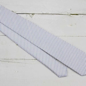 May include: A light blue and white striped necktie, diagonally positioned against a white wooden background. The tie's pattern consists of thin, parallel stripes. The fabric appears to have a subtle texture.