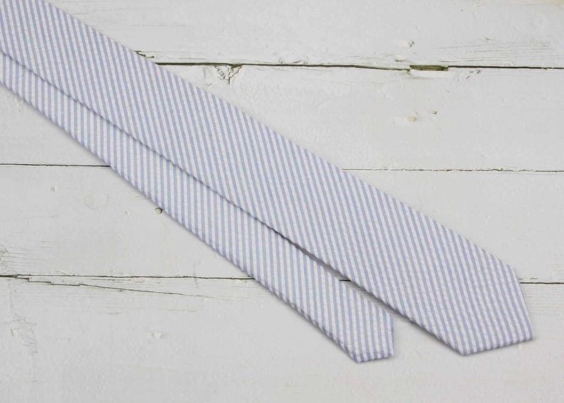 May include: A light blue and white striped necktie, diagonally positioned against a white wooden background. The tie's pattern consists of thin, parallel stripes. The fabric appears to have a subtle texture.