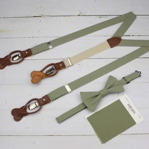 May include: A set of sage green suspenders with brown leather accents and a matching bow tie. The suspenders have adjustable straps and metal clips. The bow tie is made of the same fabric as the suspenders. The set is packaged with a fabric swatch in the same color.