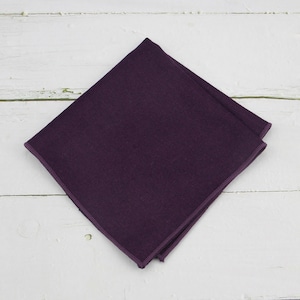 May include: A folded, square, dark purple pocket square. The fabric has a subtle texture and a darker purple border. The pocket square is displayed on a white, weathered wooden surface.