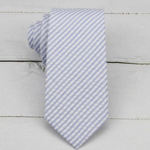 May include: A light blue and white striped necktie. The tie is made of fabric with a diagonal stripe pattern. The tie is knotted at the top and lies flat on a white wooden surface. The tie is a classic accessory for formal or business attire.