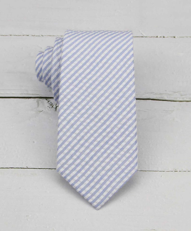 May include: A light blue and white striped necktie. The tie is made of fabric with a diagonal stripe pattern. The tie is knotted at the top and lies flat on a white wooden surface. The tie is a classic accessory for formal or business attire.