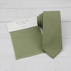 May include: A pistachio-coloured tie and fabric swatch are presented on a white wooden surface. The tie is rolled, revealing its texture and colour. The fabric swatch, labelled "AZAZIE Chiffon/Lining Pistachio", matches the tie's shade, suggesting a coordinated set.