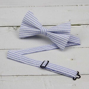 May include: A light blue and white striped bow tie with an adjustable strap and black hardware. The bow tie is made of fabric and is laying on a white wooden surface.