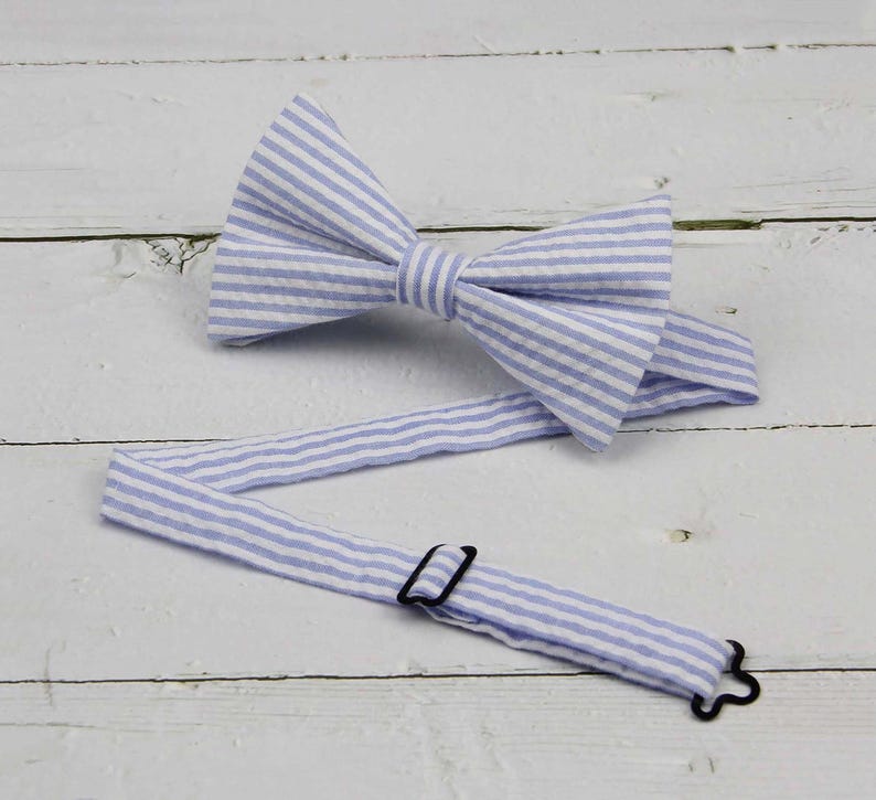 May include: A light blue and white striped bow tie with an adjustable strap and black hardware. The bow tie is made of fabric and is laying on a white wooden surface.