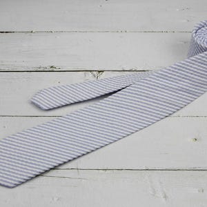 May include: A light blue and white striped tie, partially rolled, lies on a white wooden surface. The tie is made of a textured fabric with a classic seersucker pattern. The tie is a standard length.