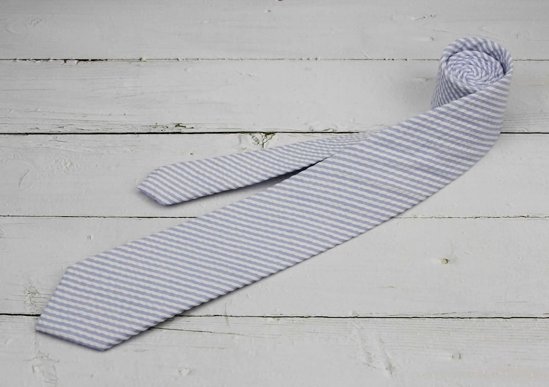 May include: A light blue and white striped tie, partially rolled, lies on a white wooden surface. The tie is made of a textured fabric with a classic seersucker pattern. The tie is a standard length.