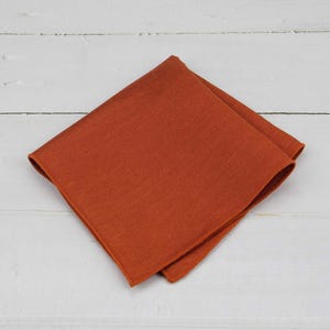 May include: A folded, square, terracotta-coloured linen napkin. The napkin is made of a textured fabric and is displayed on a white wooden surface. The napkin is a solid colour and has a simple, elegant design.