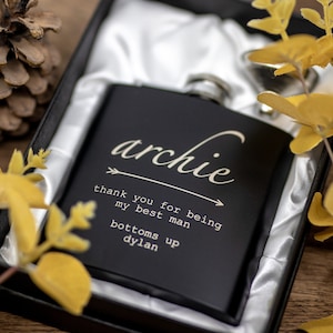 May include: Black flask with a silver lid in a black box with a white satin lining. The flask is engraved with the text "archie thank you for being my best man bottoms up dylan".