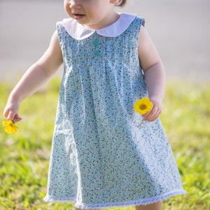 Könnte beinhalten: Ein kleines Kind trägt ein hellblaues Kleid mit weißem Kragen und Blumenmuster und hält eine gelbe Blume. Das Kleid hat einen weißen Spitzenbesatz und das Kind trägt weiße Socken und weiße Schuhe mit rosa Akzenten.