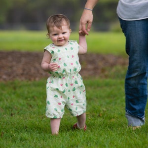 May include: A baby wearing a light green romper with a pattern of pink and green butterflies, standing on green grass. The baby is holding an adult's hand and wearing red shoes.