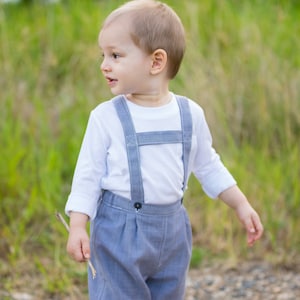 Peut inclure: Un jeune garçon portant une chemise blanche à manches longues avec des bretelles et un short à carreaux bleus et blancs. Il tient un petit bâton dans sa main droite.
