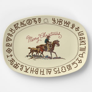 以下が含まれることがあります： 茶色のロープの縁取りが施された白い楕円形のサービングプレート。カウボーイが子牛を連れて馬に乗っている。赤い筆記体で「メリークリスマス」と書かれている。縁取りには様々なシンボルと文字が飾られている