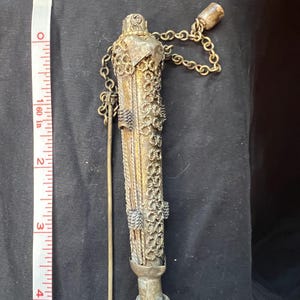 May include: An antique silver-toned writing instrument, approximately 4 inches long, with intricate filigree and chain details. The pen has a decorative top and a small, round base. A measuring tape is next to the pen.