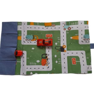 May include: A rolled-up, green and white road map play mat with a blue denim pocket and two red toy cars. The mat features a road design with trees, trucks, and the word "EXPLORE".