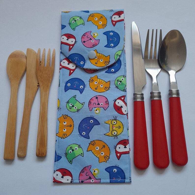 May include: A set of reusable cutlery with a blue and white fabric case featuring a repeating pattern of colorful cats. The set includes a wooden spoon, fork, and knife, and a red handled knife, fork, and spoon.