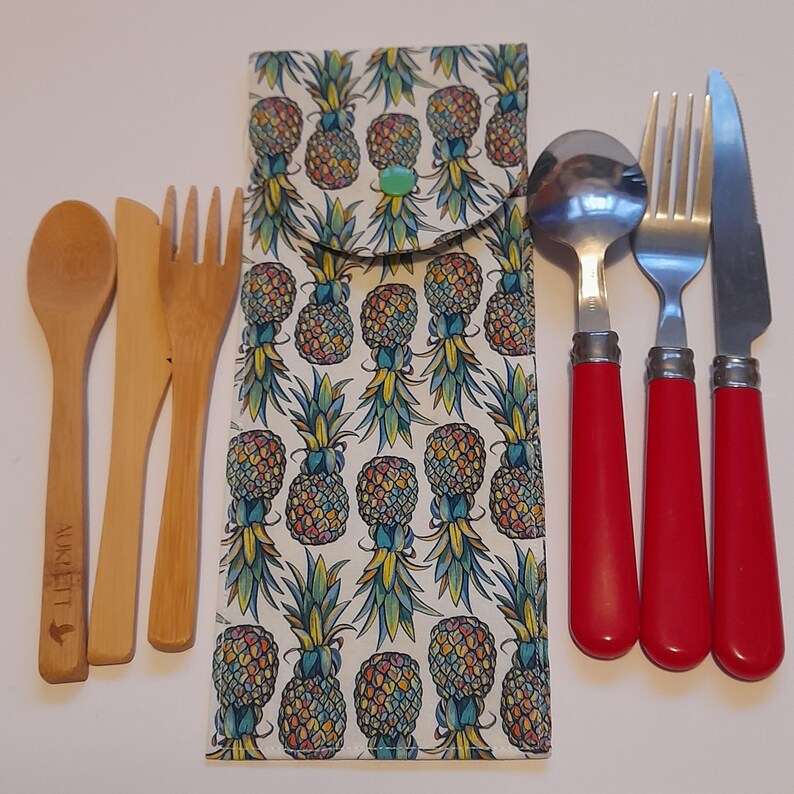 May include: A set of reusable cutlery in a pineapple patterned fabric pouch. The set includes a spoon, fork, knife, and a small wooden knife. The cutlery is made of stainless steel and has red handles. The pouch is made of cotton and has a zipper closure.