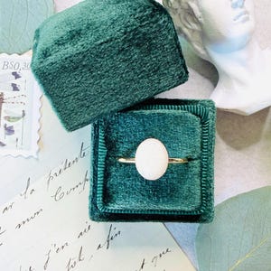 Puede incluir: Un anillo de oro con una piedra blanca redonda y grande se muestra en un joyero de terciopelo verde esmeralda abierto. La caja se encuentra sobre una superficie con escritura manuscrita y una pequeña escultura de busto blanco en el fondo.