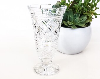 May include: A clear glass vase with a cut glass design. The vase has a wide mouth and a narrow base. It is sitting on a white surface.