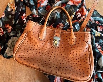 Puede incluir: Un bolso de mano de cuero marrón con una superficie texturizada y un asa superior descansa sobre un vestido negro con estampado floral. El vestido presenta rosas rojas, blancas y amarillas. El bolso tiene un cierre plateado.