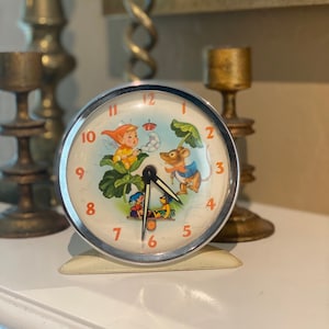 May include: A vintage alarm clock with a white face and black hands. The clock face features a colorful illustration of a fairy, a mouse, and two children. The clock has a silver metal case and a white plastic base.