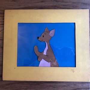 Puede incluir: Una animaci&oacute;n de celuloide enmarcada de un personaje de canguro marr&oacute;n y blanco sobre un fondo azul. El personaje mira hacia la izquierda y tiene una expresi&oacute;n de sorpresa.