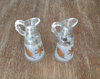 Puede incluir: Dos jarras de vidrio transparente para aceite y vinagre con diseños de hojas de oro. Las jarras tienen un acabado de vidrio esmerilado y tienen forma de jarras pequeñas.