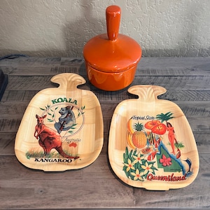 May include: Two wooden serving trays with a woven bamboo design. One tray features a kangaroo and a koala with the text "KOALA" and "KANGAROO". The other tray features a pineapple, bananas, flowers, and a woman with the text "Tropical State" and "Queensland".