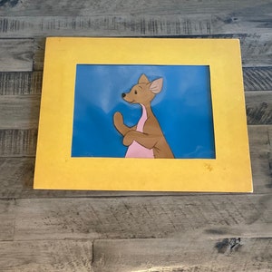 Puede incluir: Una ilustraci&oacute;n enmarcada de un canguro de dibujos animados con la barriga rosa, de pie sobre un fondo azul. El marco es amarillo.