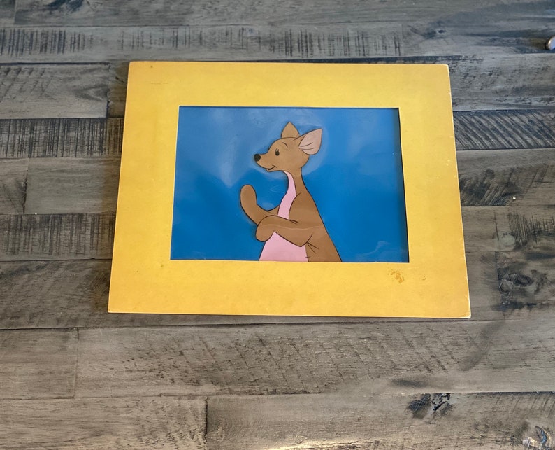 Puede incluir: Una ilustraci&oacute;n enmarcada de un canguro de dibujos animados con la barriga rosa, de pie sobre un fondo azul. El marco es amarillo.