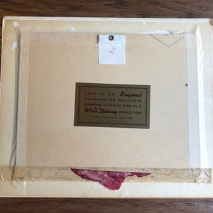 Puede incluir: Un primer plano de la parte posterior de una obra de arte enmarcada. La obra de arte es un celuloide pintado a mano de una producci&oacute;n de Walt Disney. La etiqueta dice "Este es un celuloide original pintado a mano, dibujo realmente utilizado en una producci&oacute;n de Walt Disney."