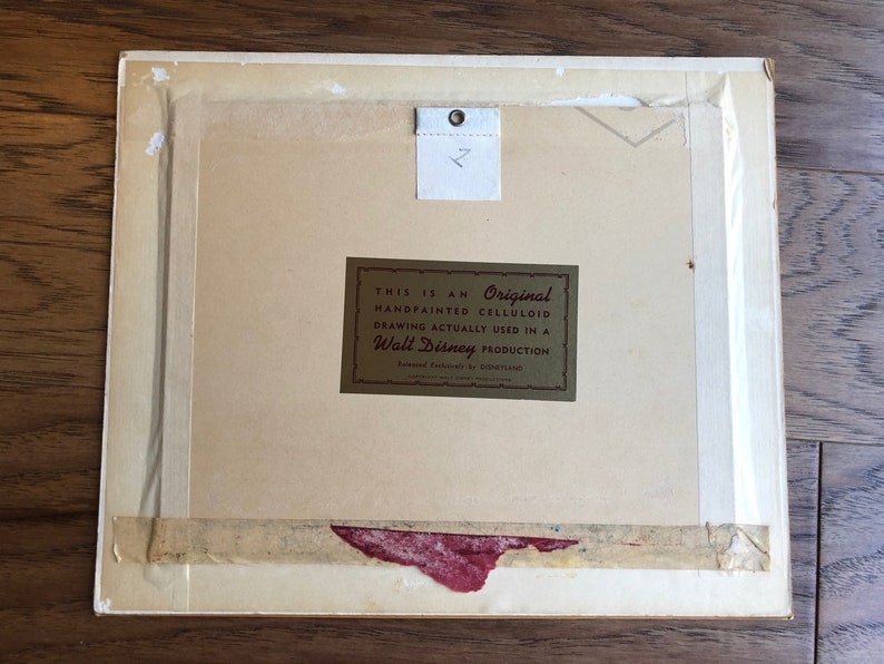 Puede incluir: Un primer plano de la parte posterior de una obra de arte enmarcada. La obra de arte es un celuloide pintado a mano de una producci&oacute;n de Walt Disney. La etiqueta dice "Este es un celuloide original pintado a mano, dibujo realmente utilizado en una producci&oacute;n de Walt Disney."