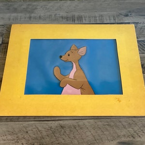 Puede incluir: Un cel de dibujos animados enmarcado de un canguro marr&oacute;n y rosa con un fondo azul. El marco es amarillo.