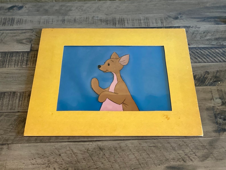 Puede incluir: Un cel de dibujos animados enmarcado de un canguro marr&oacute;n y rosa con un fondo azul. El marco es amarillo.