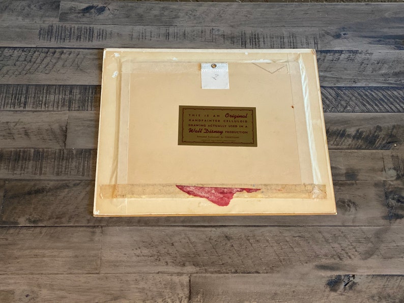 Puede incluir: Un marco blanco con un passe-partout y una etiqueta dorada que dice "This is an Original Hand Painted Celluloid Drawing Actually Used in a Walt Disney Production".