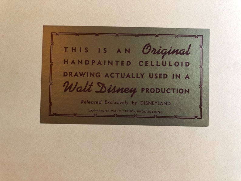 Puede incluir: Una etiqueta dorada con un borde burdeos y un texto que dice "THIS IS AN Original HANDPAINTED CELLULOID DRAWING ACTUALLY USED IN A Walt Disney PRODUCTION Released Exclusively by DISNEYLAND COPYRIGHT WALT DISNEY PRODUCTIONS".