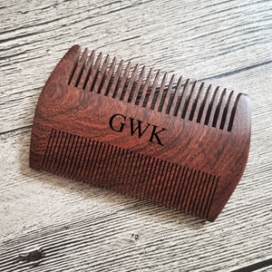 May include: A wooden beard comb with a dark reddish-brown finish. The comb has fine teeth and a curved handle. The initials "GWK" are engraved on the comb. The comb is approximately 4 inches long.