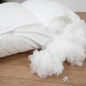 May include: A white rectangular pillow with a zippered opening along the side. The zipper is partially open, revealing a fluffy, white synthetic fiber fill that is spilling out from the interior of the pillow casing onto the surface below.
