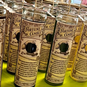 May include: A row of fortune candles with glass jars and labels that say "Fortune Candle". The labels feature a hand holding a crystal ball with a black interior. The candles are in various colors, including red, yellow, green, and brown.
