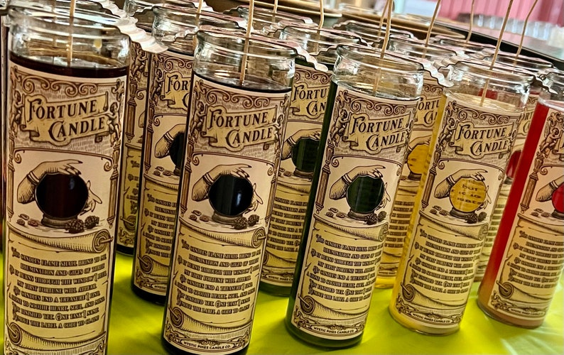 May include: A row of fortune candles with glass jars and labels that say "Fortune Candle". The labels feature a hand holding a crystal ball with a black interior. The candles are in various colors, including red, yellow, green, and brown.