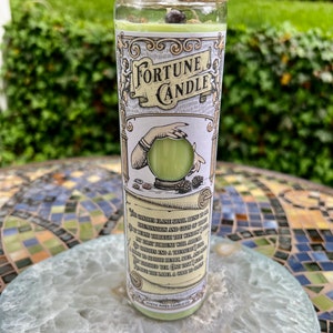 May include: A green fortune candle with a white label that reads "Fortune Candle". The label also includes a hand holding a crystal ball and the text "This candle's flame shall bring to thee illumination and gifts of truth. As it burns through the windings of my first fortune will appear. This candle with the ancient art is bound to end a treasure, and to soothe the heart, soul, and mind. One may ask a question, then light the candle and read the label. A gift to carry."