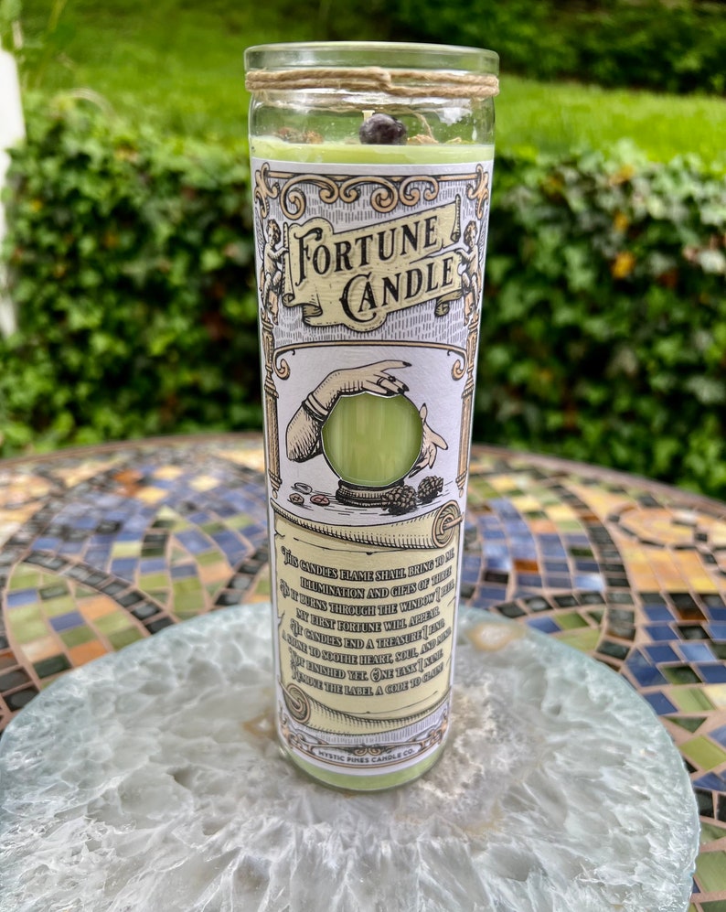 May include: A green fortune candle with a white label that reads "Fortune Candle". The label also includes a hand holding a crystal ball and the text "This candle's flame shall bring to thee illumination and gifts of truth. As it burns through the windings of my first fortune will appear. This candle with the ancient art is bound to end a treasure, and to soothe the heart, soul, and mind. One may ask a question, then light the candle and read the label. A gift to carry."