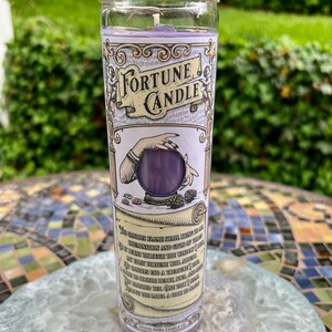 May include: A purple fortune candle in a glass jar with a twine lid. The label features a hand holding a crystal ball and the text "Fortune Candle". The text on the label reads "This candle's flame shall bring to thee illumination and gifts of the..."