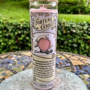 May include: A pink fortune candle with a glass jar and a twine lid. The label features a vintage illustration of a hand holding a crystal ball and the text "Fortune Candle". The text on the label reads: "This candle's flame shall bring to thee illumination and gifts of truth. If it burns through the window of thy mind, thy first fortune will appear. Let candles end a treasure, and bring light to both heart, soul, and mind. When finished, one tank to shine,  another paper, a guide to thine." Mystic Pines Candle Co.