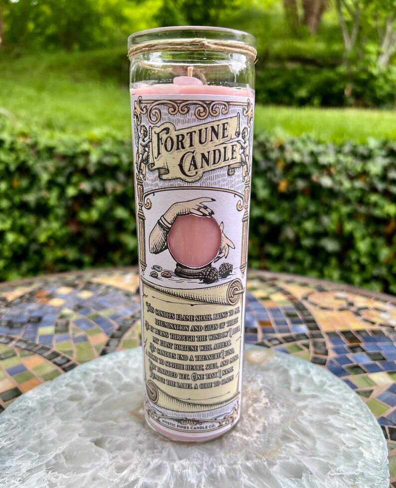 May include: A pink fortune candle with a glass jar and a twine lid. The label features a vintage illustration of a hand holding a crystal ball and the text "Fortune Candle". The text on the label reads: "This candle's flame shall bring to thee illumination and gifts of truth. If it burns through the window of thy mind, thy first fortune will appear. Let candles end a treasure, and bring light to both heart, soul, and mind. When finished, one tank to shine,  another paper, a guide to thine." Mystic Pines Candle Co.