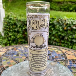 May include: A fortune candle in a clear glass jar with a white label. The label features a vintage illustration of a hand holding a crystal ball and the text "Fortune Candle". The candle is surrounded by a decorative scroll with text about the candle's purpose.