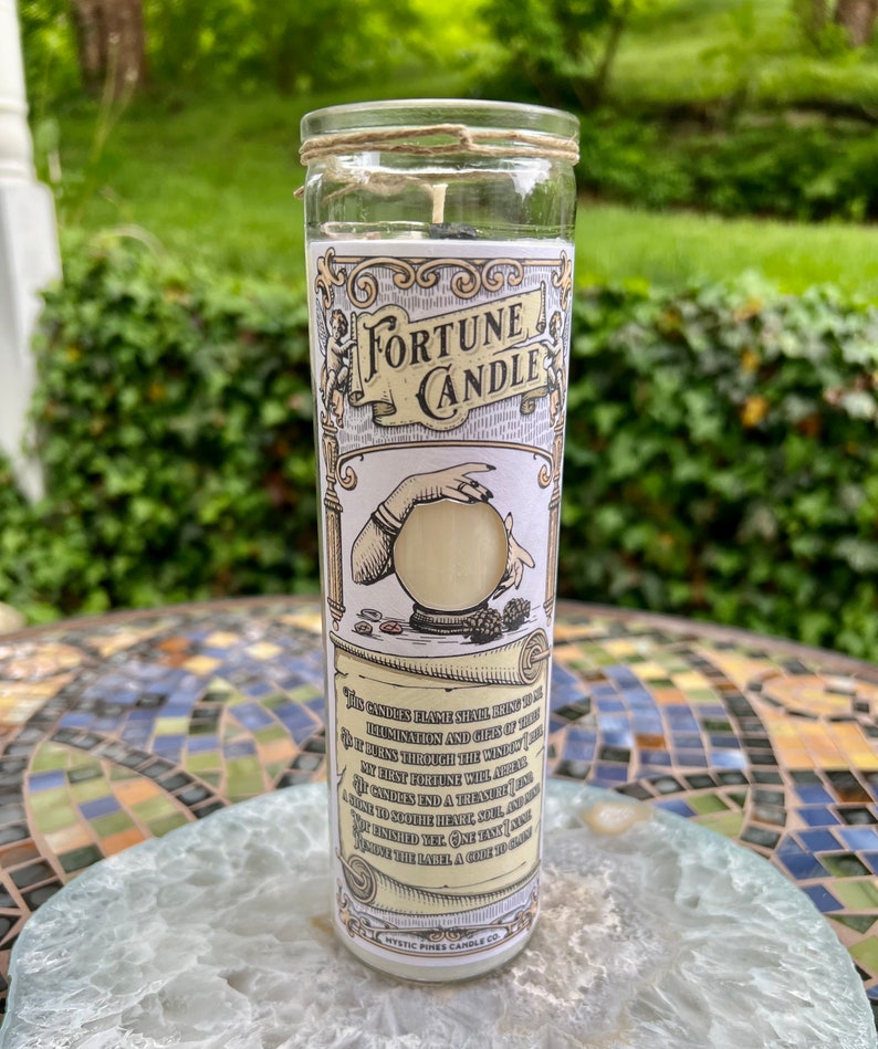 May include: A fortune candle in a clear glass jar with a white label. The label features a vintage illustration of a hand holding a crystal ball and the text "Fortune Candle". The candle is surrounded by a decorative scroll with text about the candle's purpose.