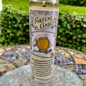 May include: A fortune candle with a yellow wax and a crystal embedded in the top. The candle is in a glass jar with a label that says "Fortune Candle" and has a quote about the candle's flame bringing hope, illumination, and gifts of their dreams. The label also says "Mystic Pines Candle Co."